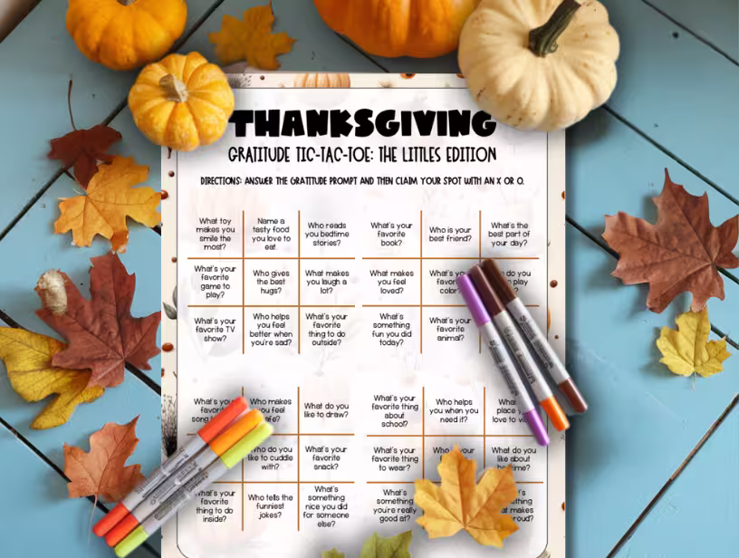 A-flat-lay-of-a-Thanksgiving-Gratitude-Tic-Tac-Toe-Game-for-Kids-titled-"The-Littles-Edition"-with-colorful-markers-and-pumpkins-on-a-blue-table-surrounded-by-autumn-leaves.